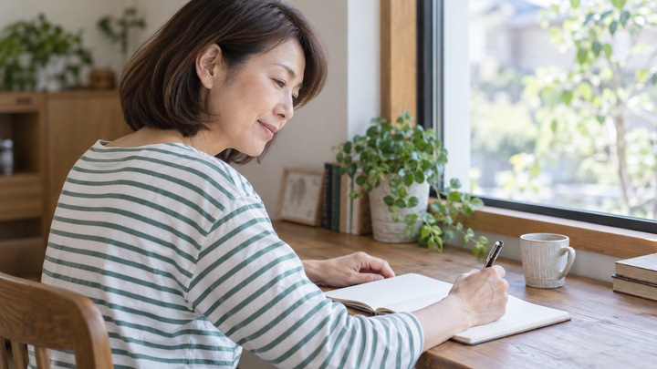 窓辺で過去の体調を振り返り、ノートに記録する日本人女性